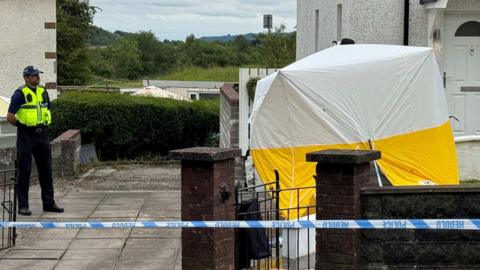 The crime scene at Rocky Road Merthyr Tydfil. There's a forensic tent outside a white house with a white garden wall. A police car is on the left hand side and there's police tape in front of the house.