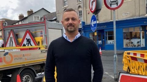 Kris Talikowski, chairman of Old Town Business Association, stood next to a road closed sign on Devizes Road in Swindon. Behind him are roadworks marked with yellow bollards and on the other side a Wales and West Utilities truck. 
