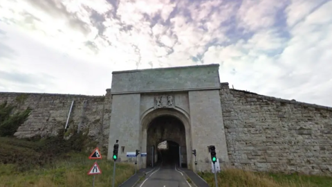 Entrance to HMP The Verne prison shows a large grey concrete arched entrance in a hillside. There are grey bricked walls either side of the entrance and traffic lights.