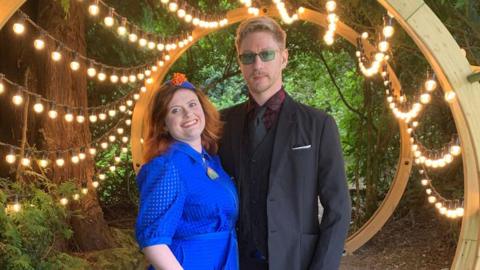 Kelly Stuart and Nick Wilson. Kelly has red hair and wears a blue dress, while Nick is blonde and wears a suit. They are surrounded by trees and a string of lightbulbs is draped around a wooden structure behind them.