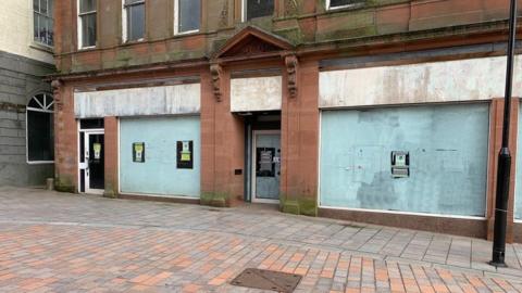 A derelict shop frontage made of sandstone with boards and the windows blanked out