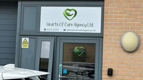 A sign above a door reads Hearts of Care Agency, listed below it are the email address and telephone number of the business. There is a green logo in the shape of a heart. On the glass door below the logo is repeated and the opening hours of the business are listed.