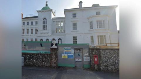 A Google Street image of a white building behind a stone wall and barriers.