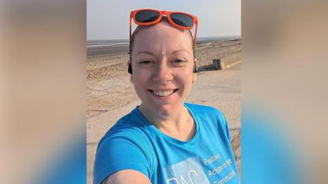 Micky Marsden with brown hair tied back and orange sunglasses resting on her head wearing a blue t-shirt with the PAC charity logo and the words Positive Action in the Community. She standing in front of a beach on a clear day. She is smiling.