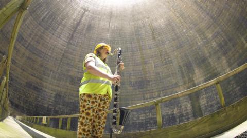 A woman wearing a hi-vis top playing holding a bass clarinet in a cooling tower