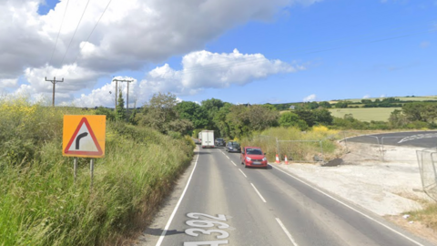 A Google Street View image of a two-lane, single carriageway section of A392, which is in a non-urban area with a few vehicles on it