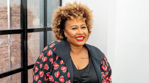 Emeli Sande smiles into the camera while standing indoors, next to a window. She is wearing a patterned jacket showing lots of hearts, and has brown hair.