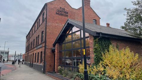 A red brick three-storey Victorian building with modern glazed front extension with pitched roof. The building is seen from the pavement, and passers-by can be seen in the distance. The Leather Museum is written on the front of the buidling.