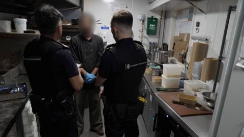 Inside a kitchen area showing the backs of two immigration officers in black uniforms, talking to a man whose face is blurred - he is wearing chef top in black and has blue food prep gloves on. Either side are stainless steel worktops with food being prepared.