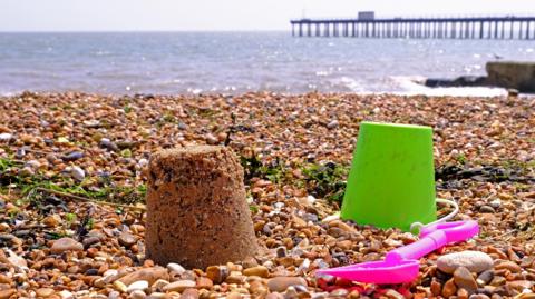 A sandcastle rests on a shingle beach. A green bucket and pink space rest next to it. A pier can be seen in the background leading into the sea.