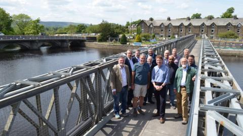 A group of people stand on a footbridge, which runs over a river, on a sunny day. In the background are some houses and another bridge.