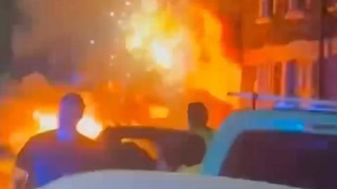 Flames and sparks appear from a residential terrace at night, with silhouetted emergency workers and vehicles in the foreground