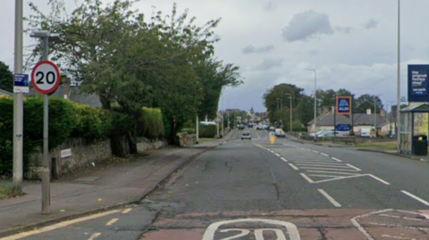 A high street with the entrance to a supermarket on one side and a trees on the other. A sign says the speed limit is 20mph