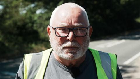 Tony Maher is wearing a grey T-shirt and a hi-vis vest. He has short grey hair, a beard and glasses. He looks serious in his expression, standing next to a road.