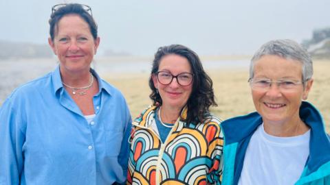 Martha Marshall-Clack, Zoe Woodward and Janet Shephard are standing on Perranporth smiling at the camera. It is a foggy day but the sane and sea can be made out behind them as well of the sides of the bay. 