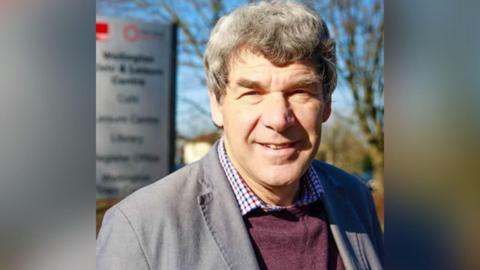 A man with short grey hair, a grey jacket, burgundy jumper and chequered shirt, stands and smiles. Behind him is a blurred out sign and a trees.