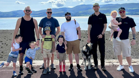 Five men stood in a row, with four young boys and a dog stood in front of them, and one man holding a young girl in his arms. They are all looking at the camera and smiling. Behind them is a body of water, with mountains in the distance.