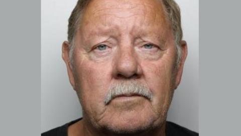 Police custody picture of a man with a grey moustache and hair. He is looking directly at the camera and is wearing a black T-shirt.