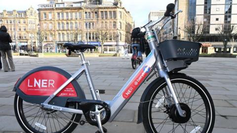 A grey and red e-bike parked in a paved city centre square on a sunny day. A man cycles away in the background