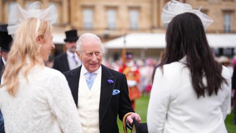 King Charles III talks with guests during a Royal Garden Party at Buckingham Palace. The King is wearing a morning suit with a pale-yellow waistcoat and light blue tie. He has short white hair and is holding a top hat in his hand. Two women have their backs to the cameras as they speak with the King. They both have long hair and are wearing white jackets and white fascinators. One has dark hair and the other one is blonde. 