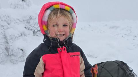 Five-year-old Faye is smiling at the camera in a snowy environment. She is wearing a bright pink jacket with black and beige sleeves. The hood is up over the top of the hood of a multi-coloured fleece. She has blonde hair which has bits of snow on it. She is standing next to a rucksack.