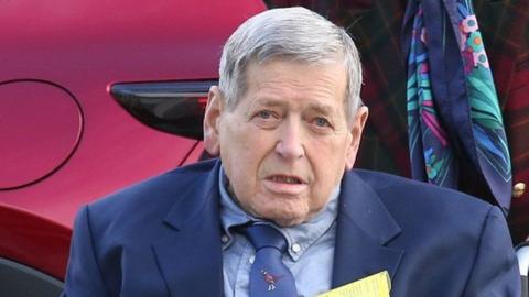 William MacDowell is an older man with grey/white hair. He is wearing a dark blue suit jacket over a light blue shirt and dark blue tie.