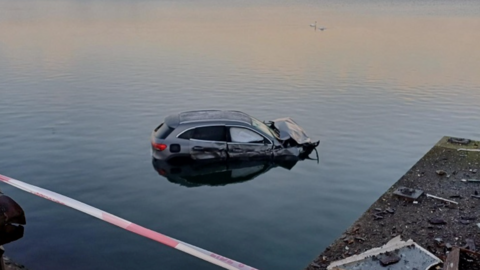 A silver Mercedes car with severe damage to its bonnet and driver's side, partially submerged in shallow water.