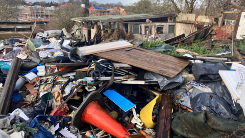 A heap of waste dumped on a piece of land. The land is strewn with a variety of rubbish including traffic cones, wooden boards and a caravan.