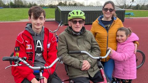 An old man sits on an adapted bike next to his 20-year-old grandson. His daughter and granddaughter are stood beside him, hugging. The man is wearing a helmet and the family are all wearing coats. They are at an athletics track.