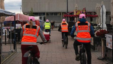 Four people in high-vis vests cycle in middle of market place and cafe area in Oulu