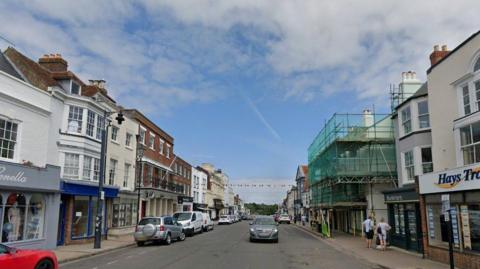 Lymington High Street shows rows of shops on either side of the street with a few cars parked on the left and hardly any parked on the right as a grey car drives down the street toward the camera. Bunting is hanging across the street in the background.