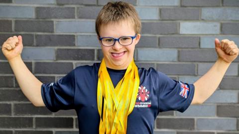 Joe Hughes, with two arms stretched out, with lots of yellow ribbons round his neck, he is smiling. He has on blue glasses and has short fair hair. He is standing by a brick wall and has on a blue T-shirt. 