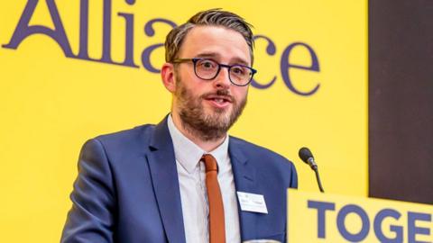 Nick Matheson has a grey beard, grey/whitish hair that is gelled into a spike and grey round glasses. He is wearing a navy shirt, white shirt and orange tie, and behind him is the yellow white Alliance banner