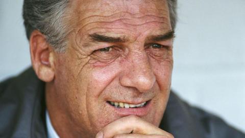 John Lyall looks on from the bench during a match in 1993. He rests his head in his hand. He wears a grey suit with a white shirt and tie. He has short grey hair. 