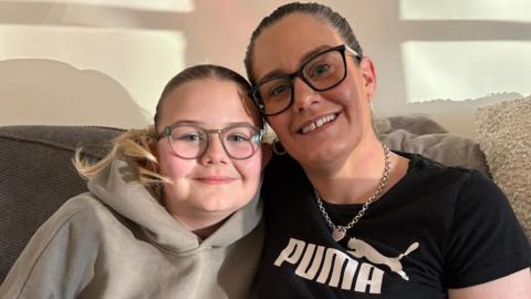 A girl with her hair in a ponytail, wearing glasses and a beige hoodie, sits on a sofa next to a woman, also with her hair back and wearing glasses, wearing a silver necklace and earrings and a Puma T-shirt with a panther on it.