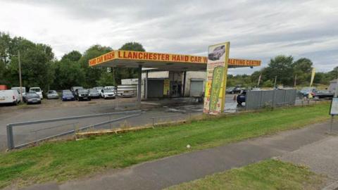 Lanchester Hand Car Wash. The car wash has been made out of a converted petrol station. It has a yellow roof with the name of the business displayed in red letters. There is an extra L in Lanchester. Rows of cars are parked behind the building.