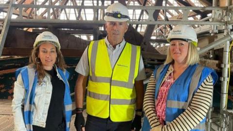 Two women are either side of one man, with all three wearing hard hats. The man is wearing a fluorescent jacket and the women are wearing blue and grey tops.