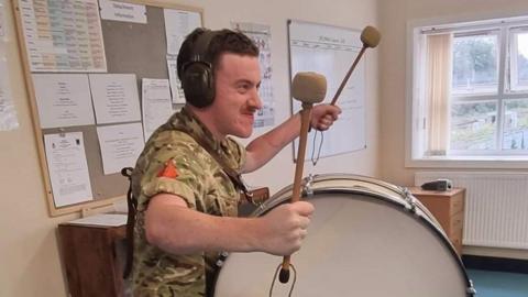 A man with short brown hair and a moustache is wearing an army combat uniform and headphones. He is standing in an office holding a pair of drumsticks and has a large drum strapped to his front. 