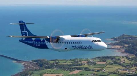 A blue and white Blue Islands aircraft flying over the Jersey countryside with the ocean in the background.