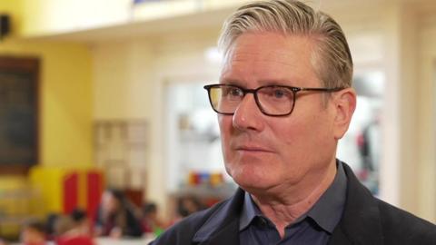 Kier Starmer, a middle-aged man with silver hair swept to the side, standing in a classroom. He is wearing black-rimmed glasses.