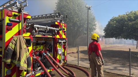 A fire engine parked on the road next to the Thomas Bullock Primary Academy building on Pound Lane in Shipdham. Hoses are attached to the back of the fire engine. A firefighter stands nearby wearing brown dungarees, a red t-shirt and a yellow helmet with visor. In the distance smoke is coming from the roof of the school building.
