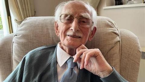 Man wearing glasses looks straight into the camera lens. He's smiling and wearing a white shirt with a blue and white spotted tie and grey cardigan