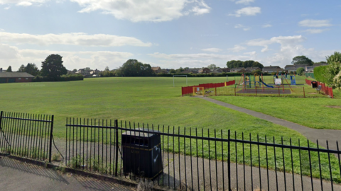 Cockton Hill Recreation Ground is a large, grassy field behind a black metal fence. There is a black bin in the foreground and a children's play park further back. In the distance to the left, there is a single storey brick building.