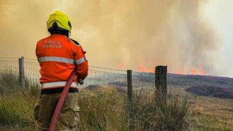 A firefighter in full kit is facing away from the camera, holding a hose which is spraying water towards greenery. Flames and a large amount of smoke are visible in the distance.