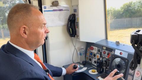 A man driving a train in the cab wearing an orange tie and blue jacket. His hands are at the controls. 