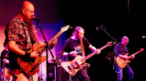 Three men holding guitars are standing on a stage that is lit up in a purple light.
