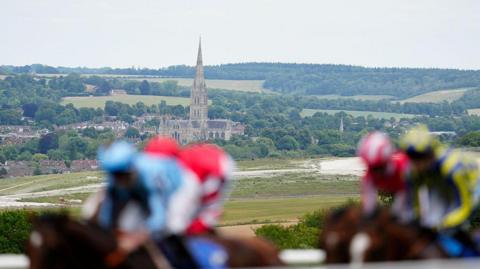 Blurred jockeys on racehorses to the front, in focus behind is green landscape with the city of Salisbury behind, the cathedral high above everything else