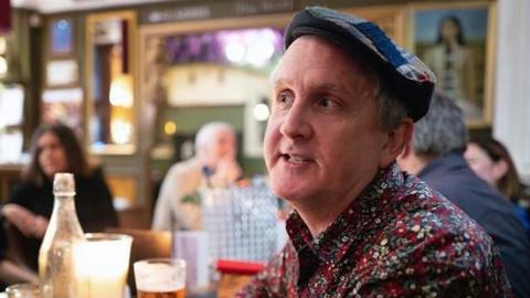 A man with grey hair is wearing a cap and a shirt with flowers on. He is sitting in a pub.