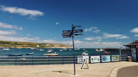 A sign post on the path next to a bay full of little boats with land in the background and blue sky above