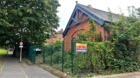 A church hall building, behind fencing which is covered with greenery. A cycle path runs past the near side of the fence.